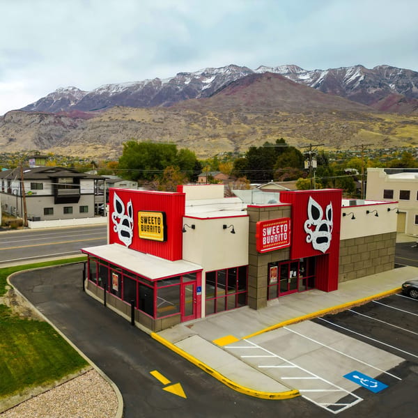 Sweeto Burrito restaurant with red facade and logo, parking lot, and snow-capped mountains in background.